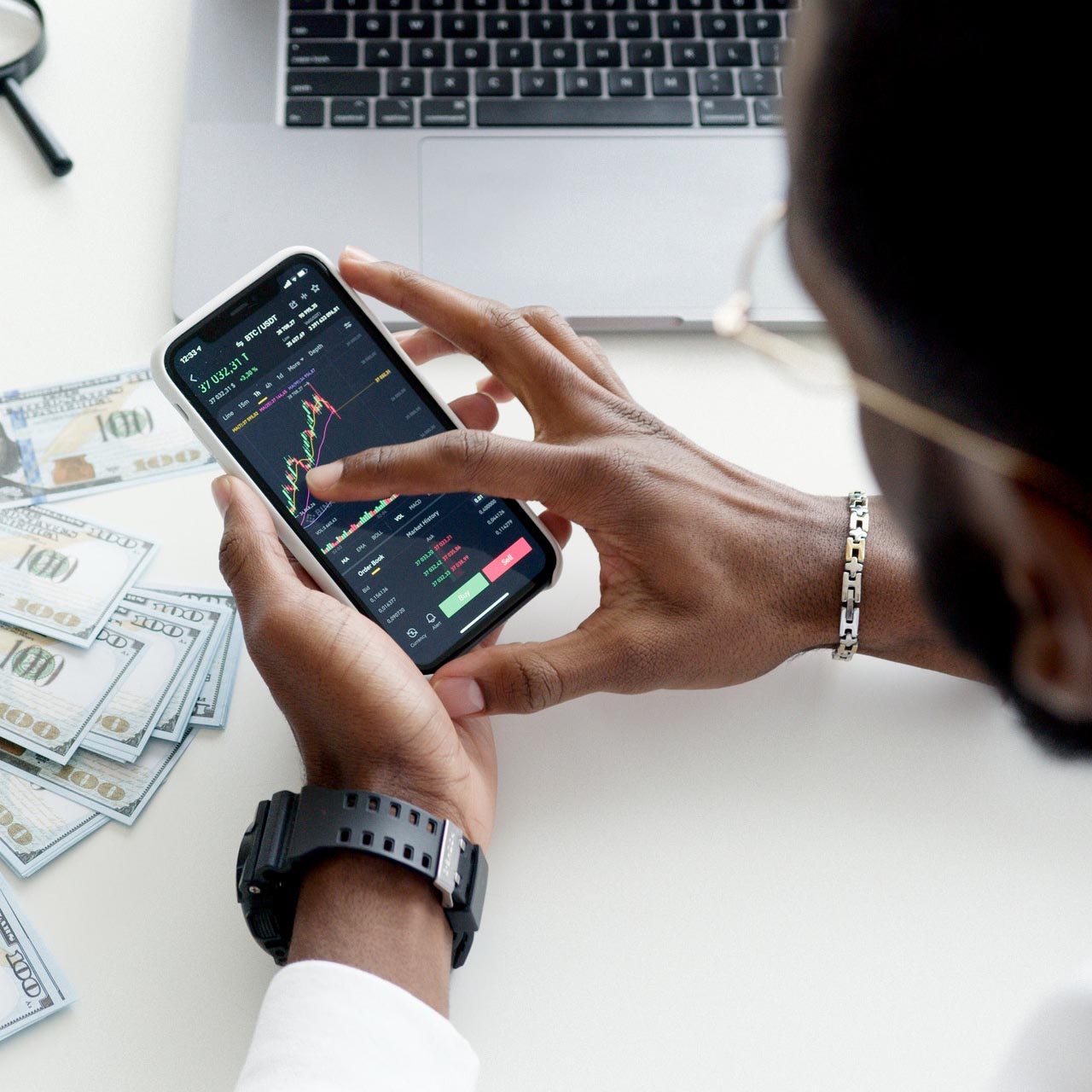 Man's hand using phone with laptop and money in the background